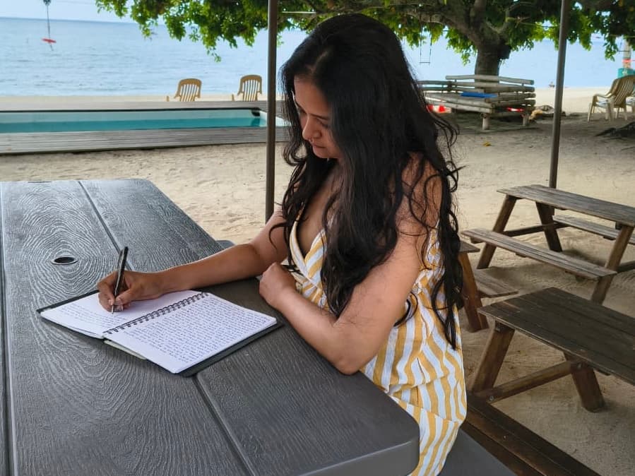 A woman with long dark hair in a yellow striped dress writes in a spiral notebook at a wooden picnic table by a sandy beach.